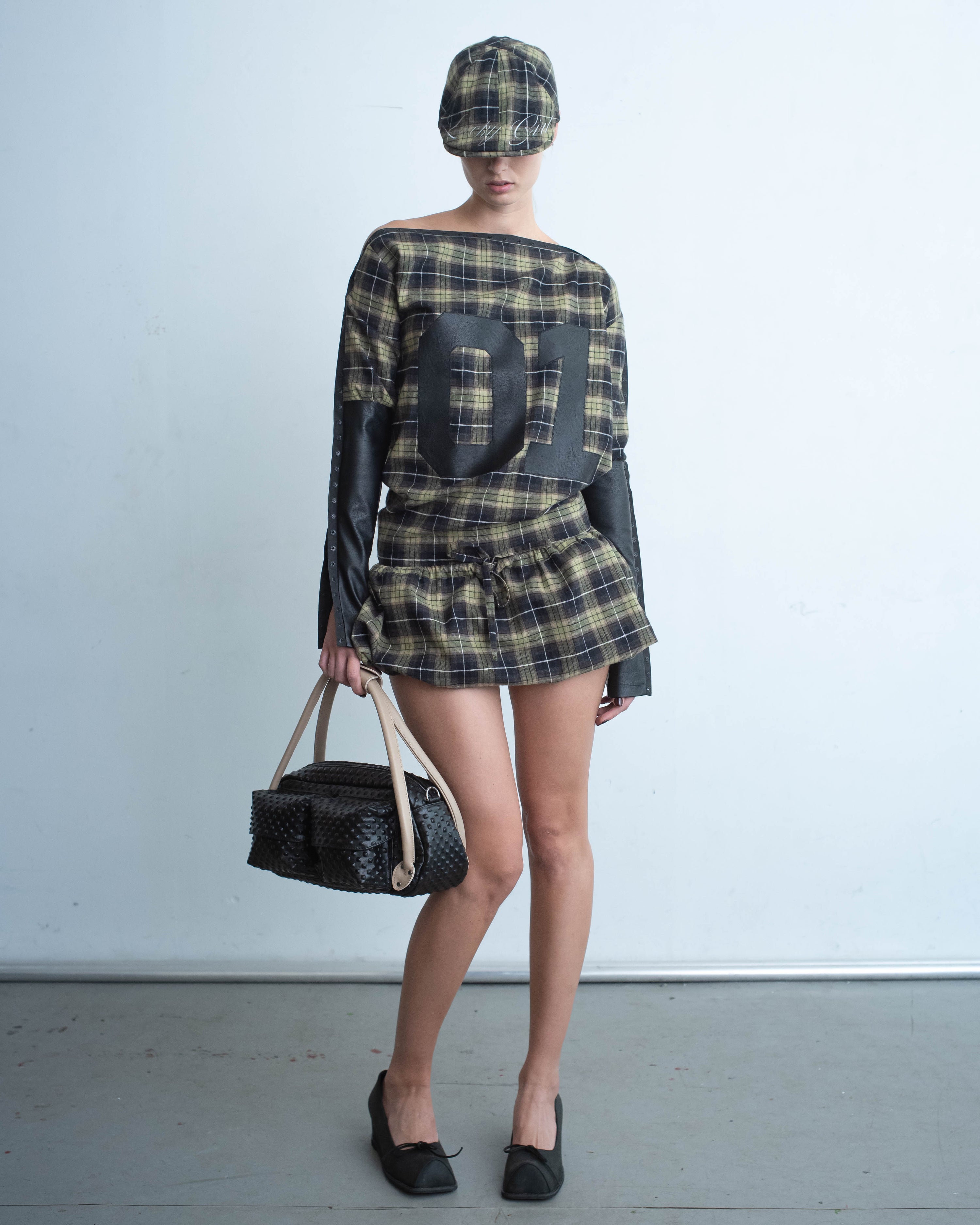A long-sleeve plaid dress with black leather paneling on the sleeves and snap details along the edges. The front features a bold "01" graphic in black, and the waist is styled cinched with a drawstring for a structured fit. 